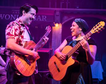 A man and a woman hold guitars and are facing each other smiling