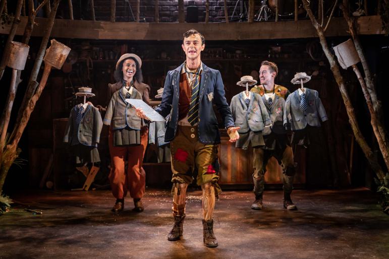 A Pinocchio production shot, with Pinocchio in a school jacket and tie holding a book and two actors with small uniforms in front of their costumes as well as holding extra uniforms to give the impression of lots of children