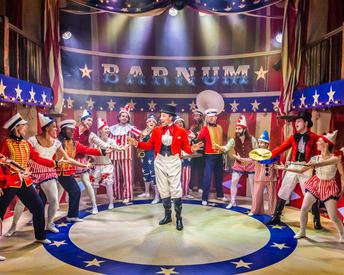 P.T Barnum standing inside a circus ring surrounded by musicians wearing a colourful marching band uniform.