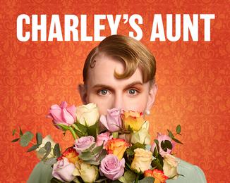 A glamourous man with perfect make up is dressed up as a woman, hiding the bottom half of him face behind some flowers.  He's on a background of patterned orange wallpaper