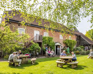 The front of Watermill Theatre in the sunshine with audiences sat on benches enjoying the sunshine.