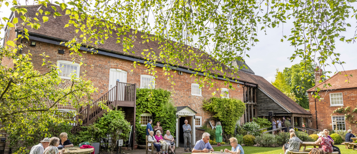The front of Watermill Theatre in the sunshine with audiences sat on benches enjoying the sunshine.