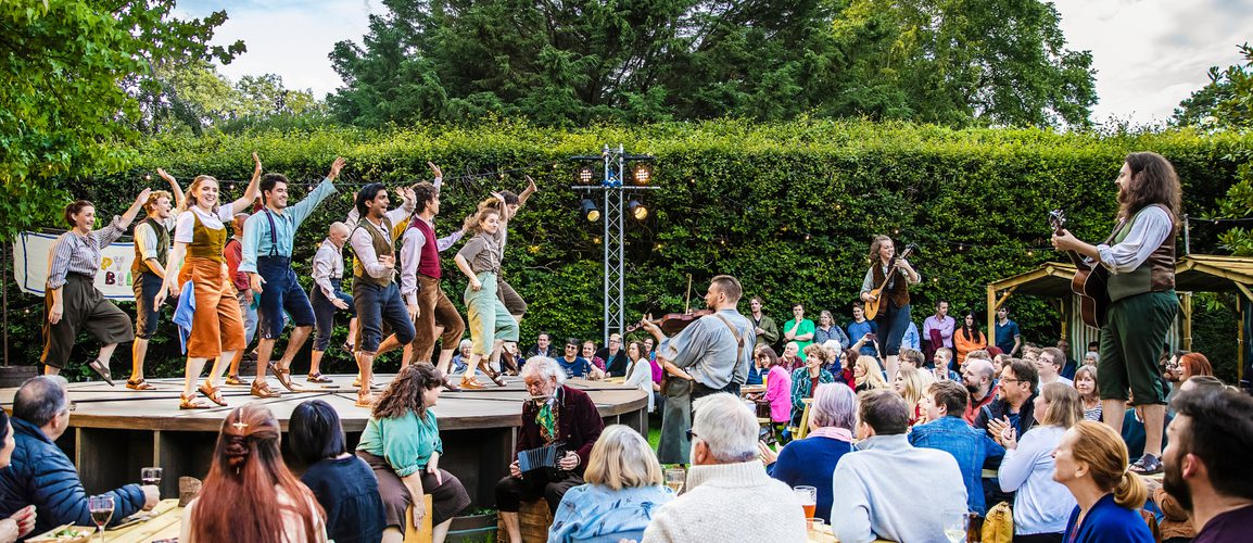 People dance on a stage outdoors, while musicians stand and play on the audiences tables.