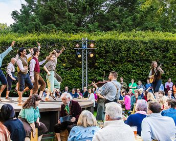 People dance on a stage outdoors, while musicians stand and play on the audiences tables.