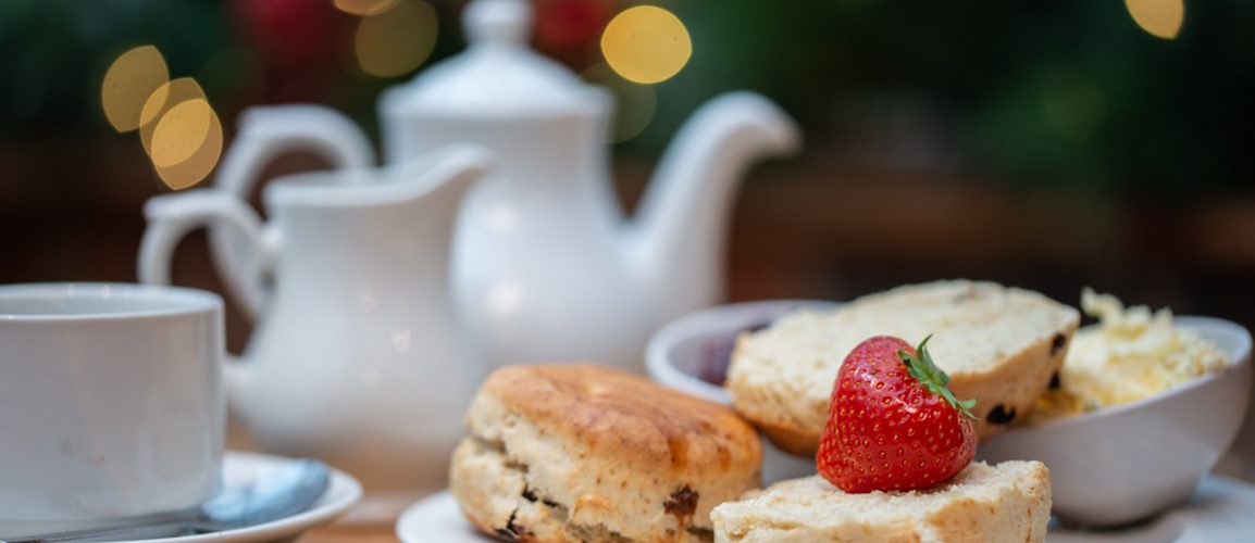 A pot of tea in a white teapot, with a plate of scones, fresh cream and jam