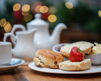 A pot of tea in a white teapot, with a plate of scones, fresh cream and jam