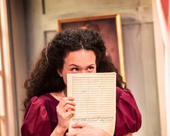A woman peeks above a handwritten music sheet with a look of mischief and excitement. She wears a period red dress.