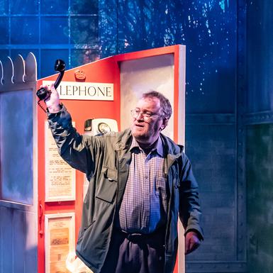 A Notes from a Small Island production photo, with Bill in a red phone box holding the phone up to the air