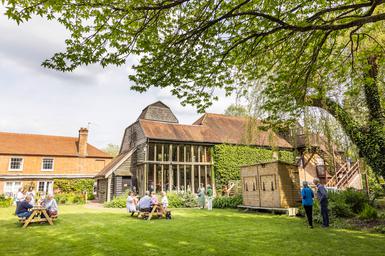 The Watermill Theatre on a sunny day with blue skies. Patrons are sat on benches on the lawn outside the venue.
