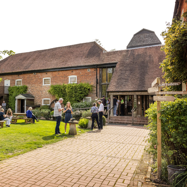 A path leading up to the Watermill Theatre with customers sat on benches on the front lawn.