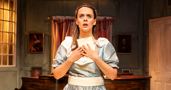 An actor in a blue dress standing in front of a piano clutching a letter.